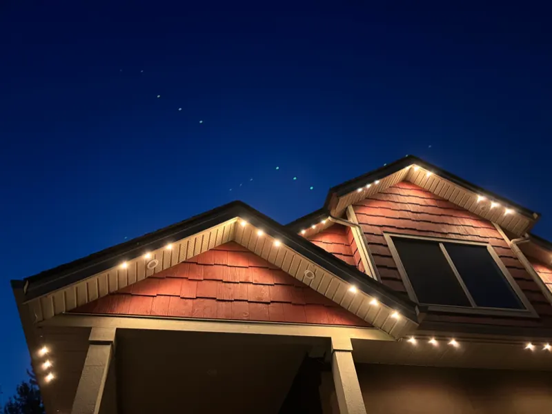 Warm white permanent lighting outlining cedar shake gables on a Castlegar home against a deep blue dusk sky