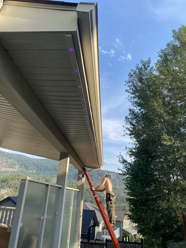 SUMMIT Lighting Systems installer mounting permanent light track from a ladder on a Castlegar BC home, Kootenay mountains behind